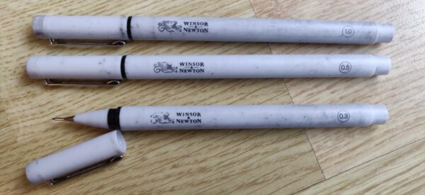 Trois crayons Winsor & Newton (tailles 1.0, 0.5 et 0.3). Le capuchon ouvert de l'un d'eux permet de voir une pointe fine, ronde, noire.