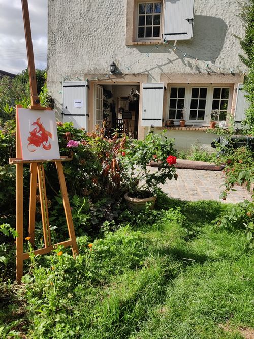 Un chevalet avec un tableau (calligraphie contemporaine quasi abstraite) avec, dans le fond, une baie vitrée permettant de rentrer dans une maison blanche