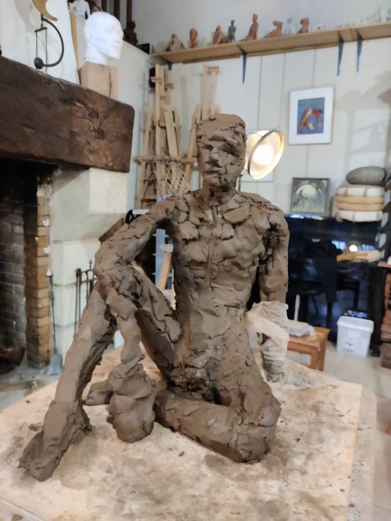 Sculpture en terre en cours de réalisation - les touches et les formes sont encore très grossières : un homme assis au sol quasiment de face, un genou replié et son coude posé dessus