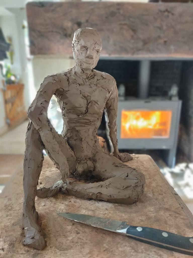 Sculpture en terre en cours de réalisation - les formes sont assez définies mais avec encore beaucoup de choses à affiner : un homme assis au sol quasiment de face, un genou replié et son coude posé dessus