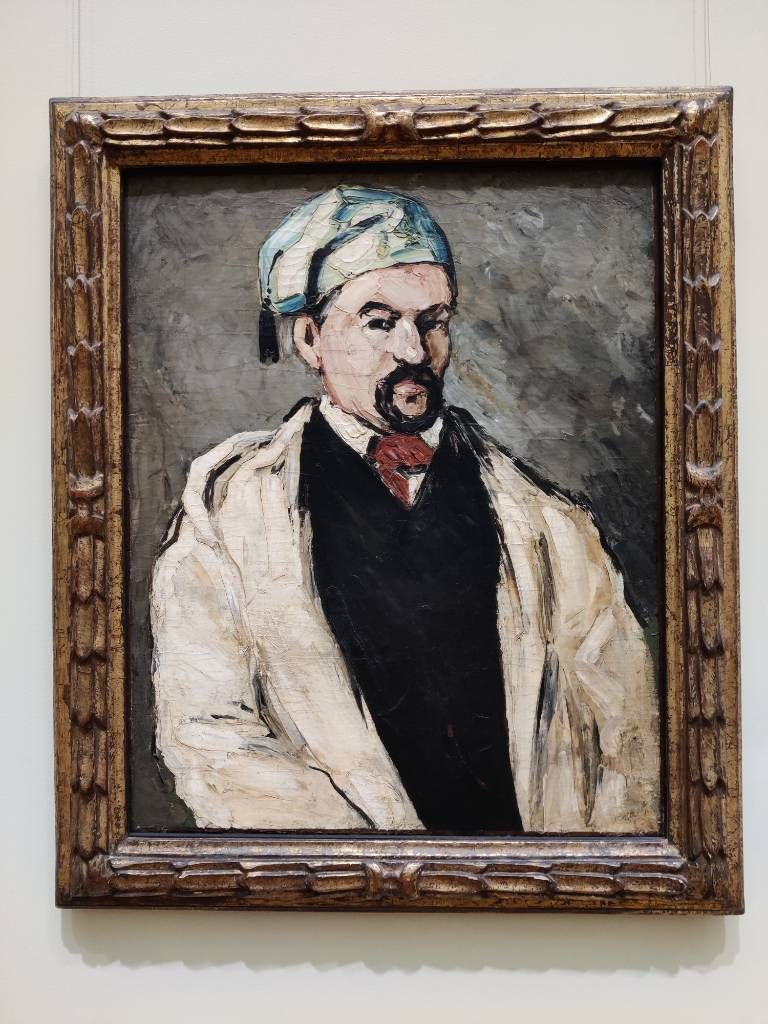 Portrait très contrasté d'un homme barbu portant une sorte de bonnet bleu, une cravate rouge, un pull noir et une chemise et un manteau blancs